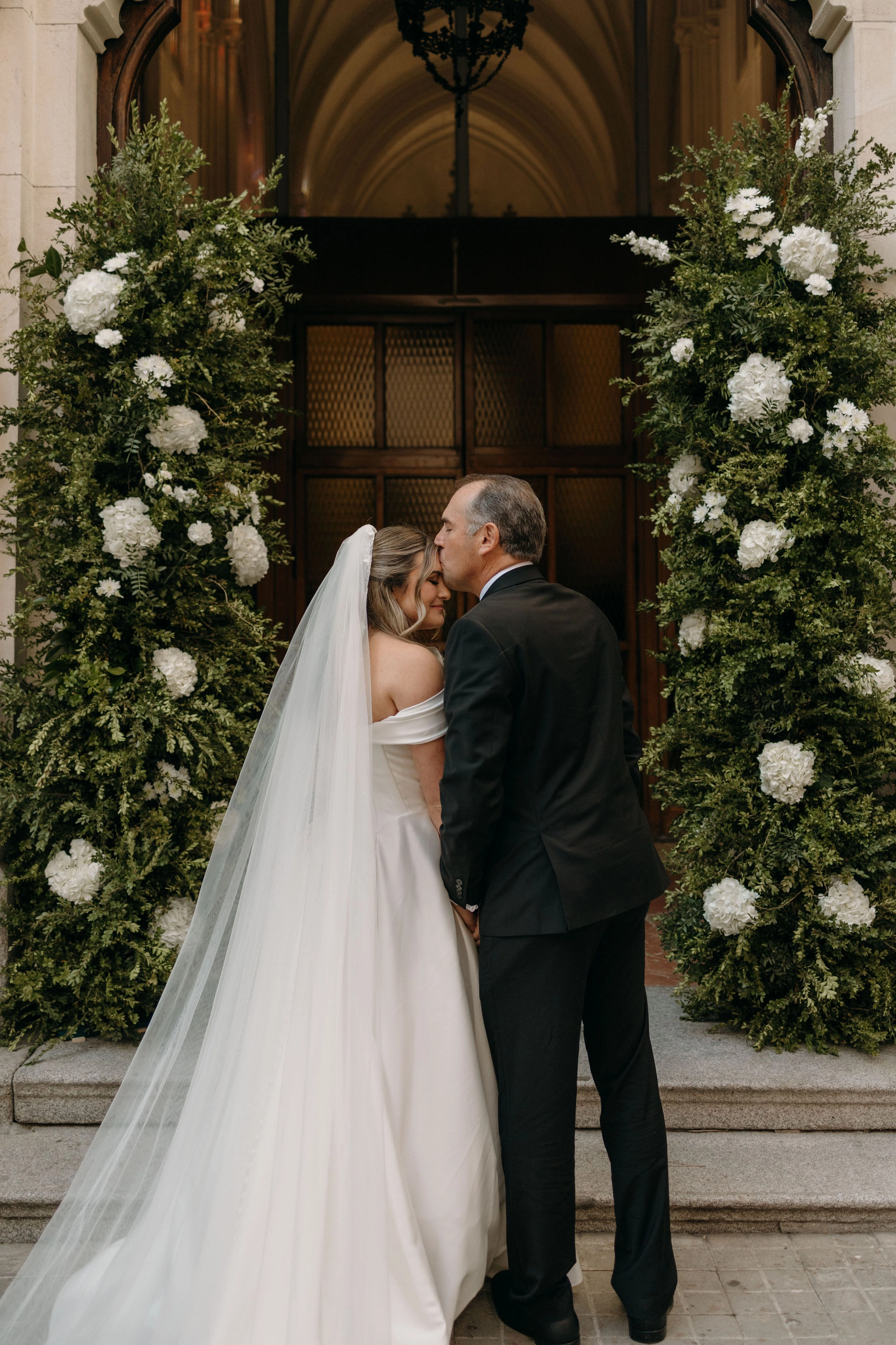 Fotografía de espaldas de una novia y su padrino frente a la entrada de una iglesia o recinto histórico.